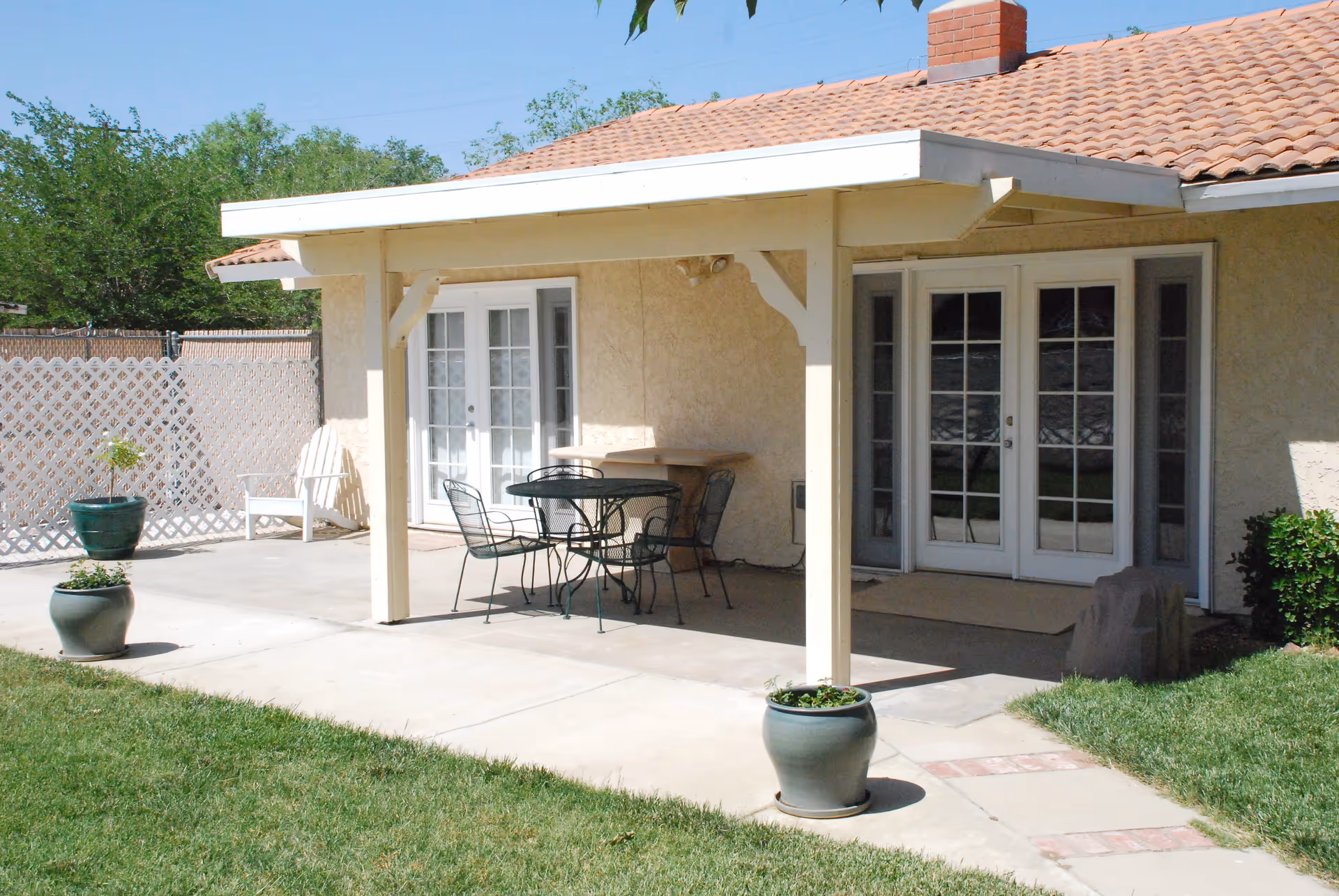 Outdoor patio area at El Vista Residential Care with a covered seating area featuring a round metal table and four chairs. The patio is surrounded by a concrete walkway and green grass, with potted plants placed around the space. The building has a beige stucco exterior and a tiled roof, with two sets of white French doors leading inside.