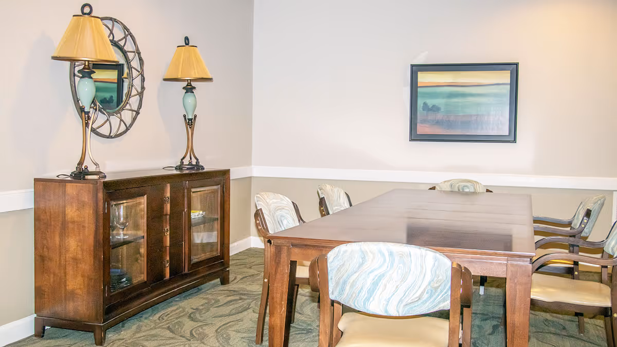 A dining/meeting room with a long wooden table surrounded by upholstered chairs, a wooden sideboard topped with two lamps and a mirror, and framed artwork on the wall.