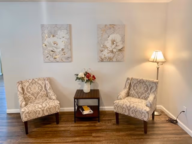 A cozy seating area with two patterned armchairs facing each other, separated by a small wooden side table holding a vase with flowers. Two floral paintings hang on the wall above the chairs, and a floor lamp stands in the corner providing warm light. The room has wooden flooring and light-colored walls.