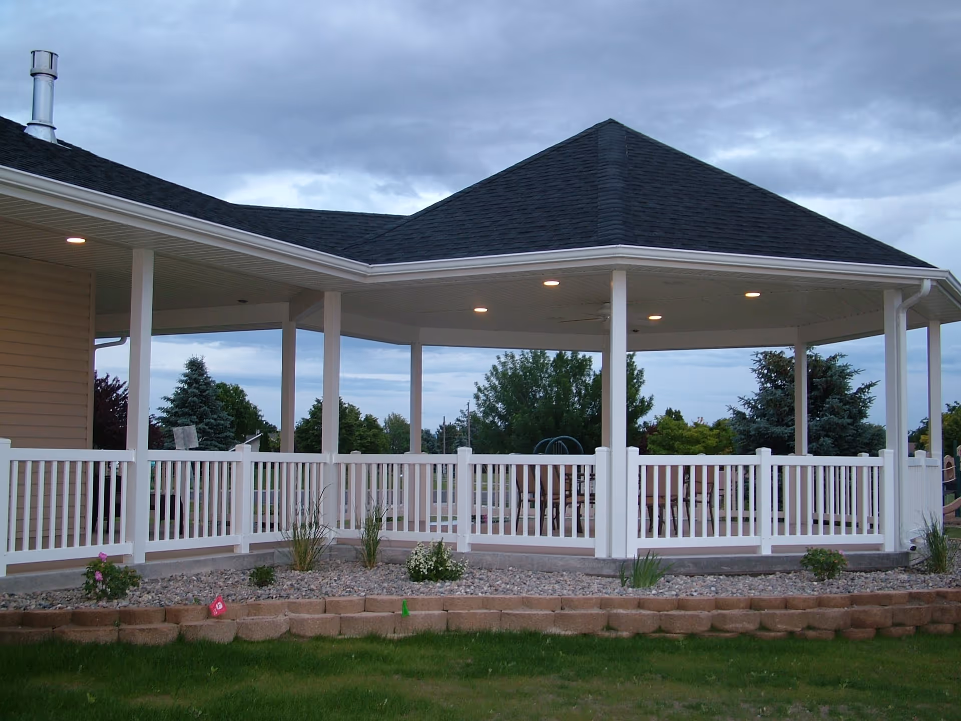 Outdoor covered patio area with white railing, stone landscaping, and green grass in front. The patio has a dark shingled roof with recessed lighting and ceiling fans. Trees and cloudy sky are visible in the background.