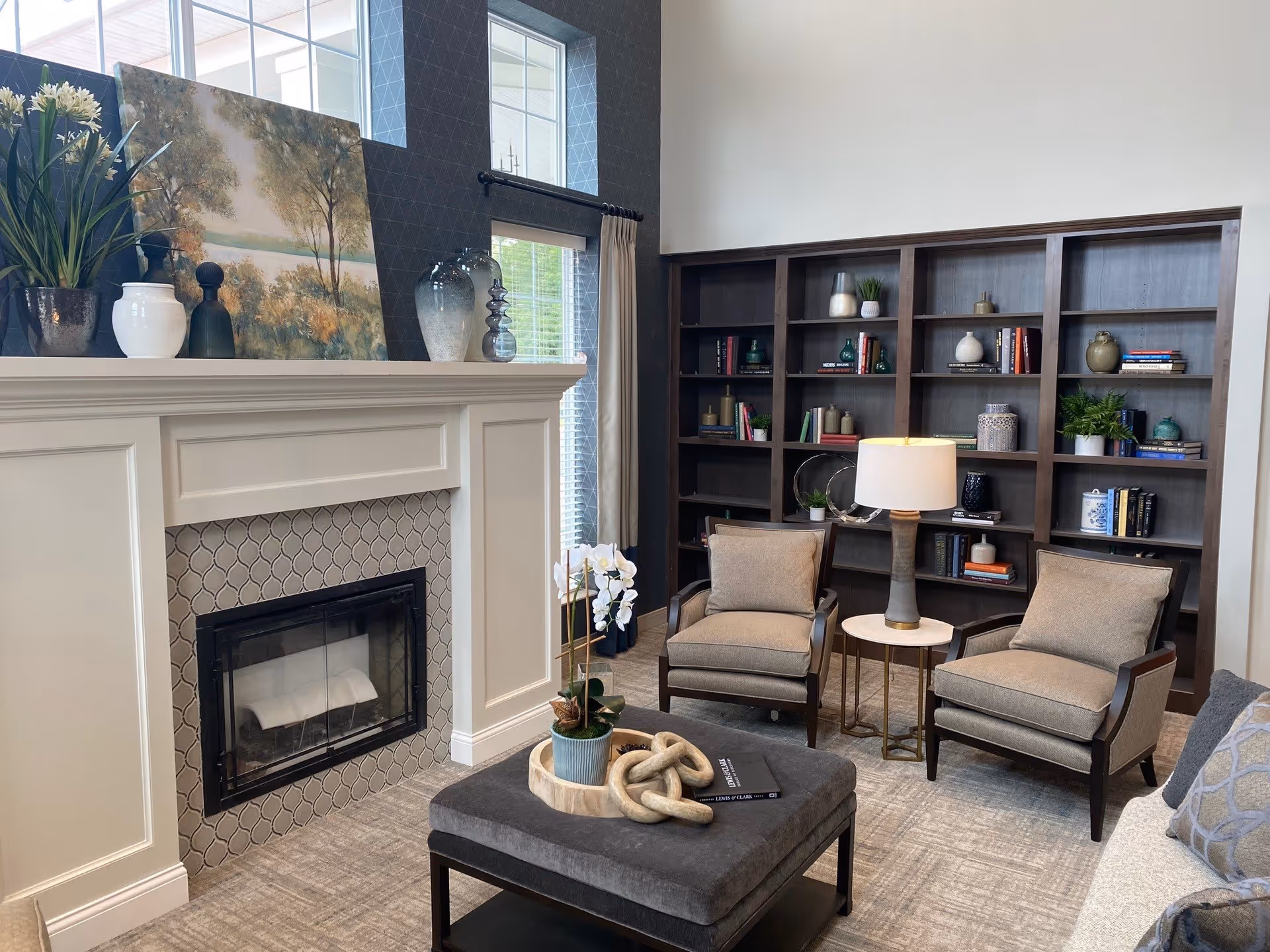 A cozy living room with a white fireplace featuring patterned tile surround, a mantel decorated with vases and a landscape painting. Two beige armchairs flank a small round table with a lamp. A large dark wood bookshelf filled with books and decorative items is against the wall. A gray ottoman with a plant and decorative objects sits in the center on a light carpeted floor.