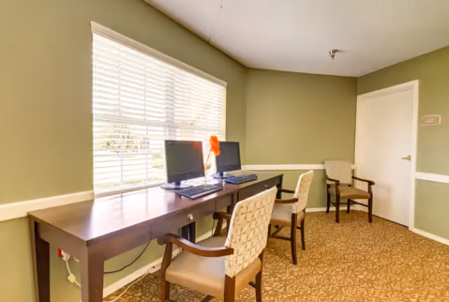 A small computer area in a senior living facility with two desktop computers on a long wooden desk. Two cushioned chairs with armrests are positioned at the desk. A window with white blinds allows natural light into the room, which has light green walls and patterned carpet. Another cushioned chair is placed near a closed white door in the corner.