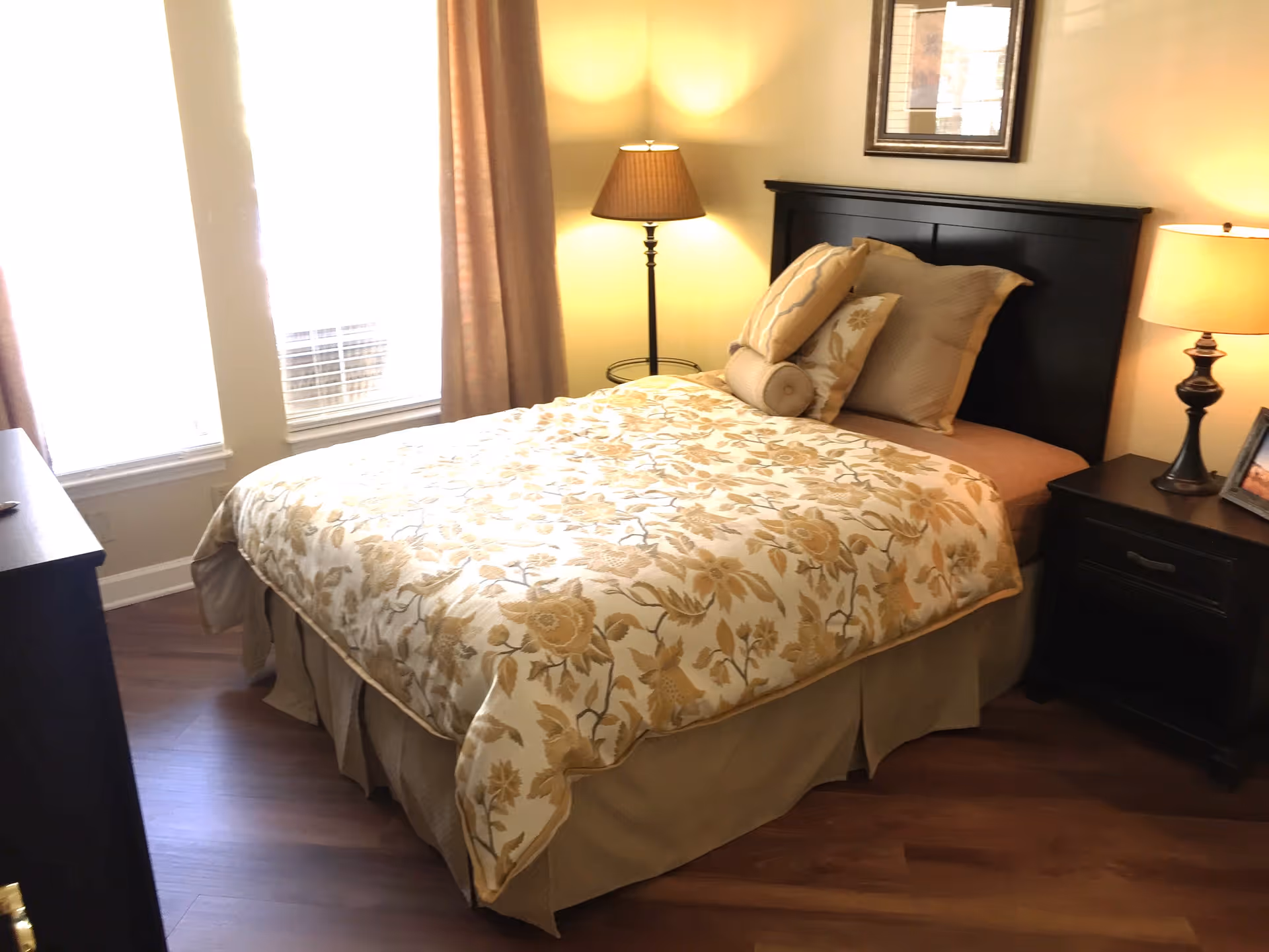 A cozy bedroom with a bed covered in a floral patterned comforter and multiple pillows. The room has two windows with beige curtains, a dark wooden headboard, two bedside tables each with a lamp, and a framed picture on the wall above the bed.