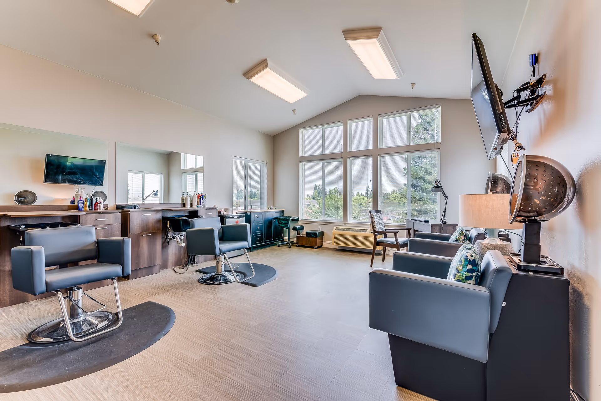 Bright salon-style common room with styling chairs, mirrors, seating and large windows overlooking greenery.