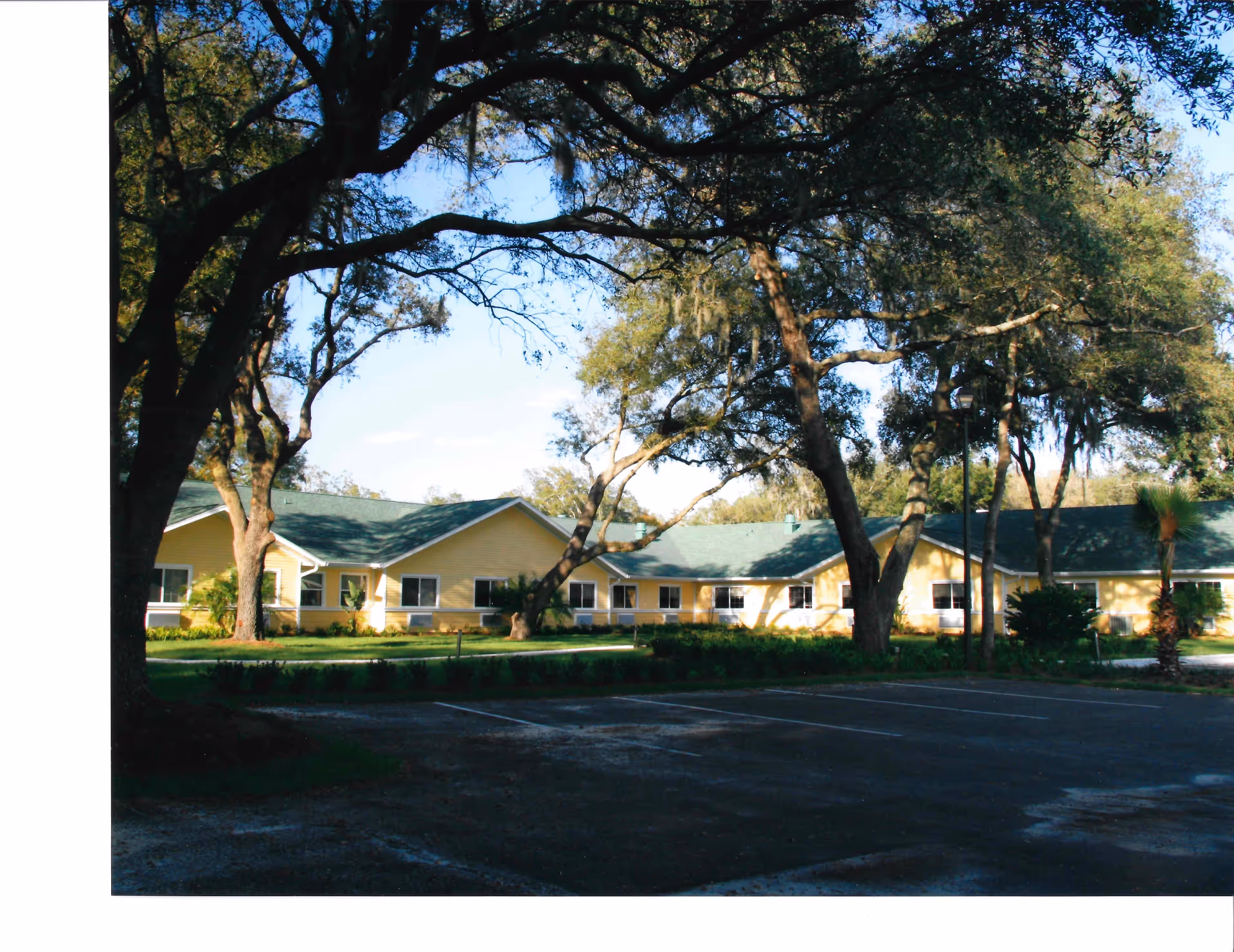 Exterior view of a single-story yellow building with a green roof surrounded by large trees and a parking lot in the foreground under a clear blue sky.