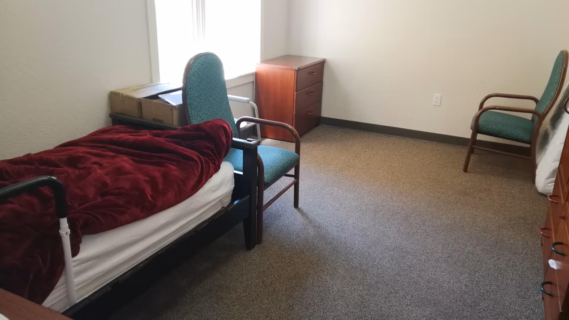 Sparsely furnished senior bedroom with a single bed covered by a red blanket, two green upholstered chairs, and a wooden dresser near a window.
