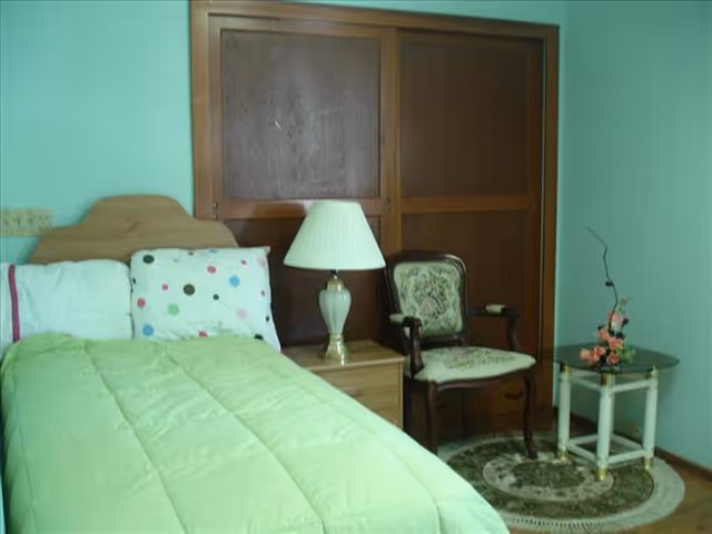 A bedroom with a single bed covered with a green comforter and a white pillow with colorful polka dots. Next to the bed is a wooden nightstand with a white lamp. There is a vintage upholstered chair and a small glass-top side table with a floral arrangement on it. The room has a wooden closet with sliding doors and light green walls.