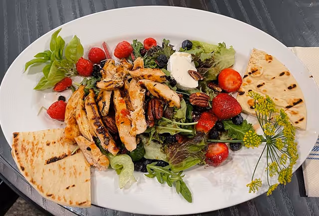 Grilled chicken salad with mixed greens, strawberries, blueberries, pecans, a dollop of cheese, flatbread, and fresh herbs on a white oval plate.