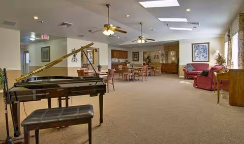 A spacious senior living common area featuring a grand piano with a bench in the foreground, several wooden tables and chairs arranged throughout the room, ceiling fans with lights, skylights, and a seating area with red upholstered chairs and a couch near a window with floral curtains.