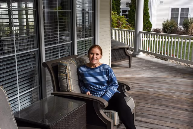A woman with red hair wearing a blue and white striped sweater and black pants is sitting on a cushioned chair on a wooden porch. The porch has white railings and columns, with another chair and a glass-top table nearby. In the background, there is a grassy area and white buildings with windows.
