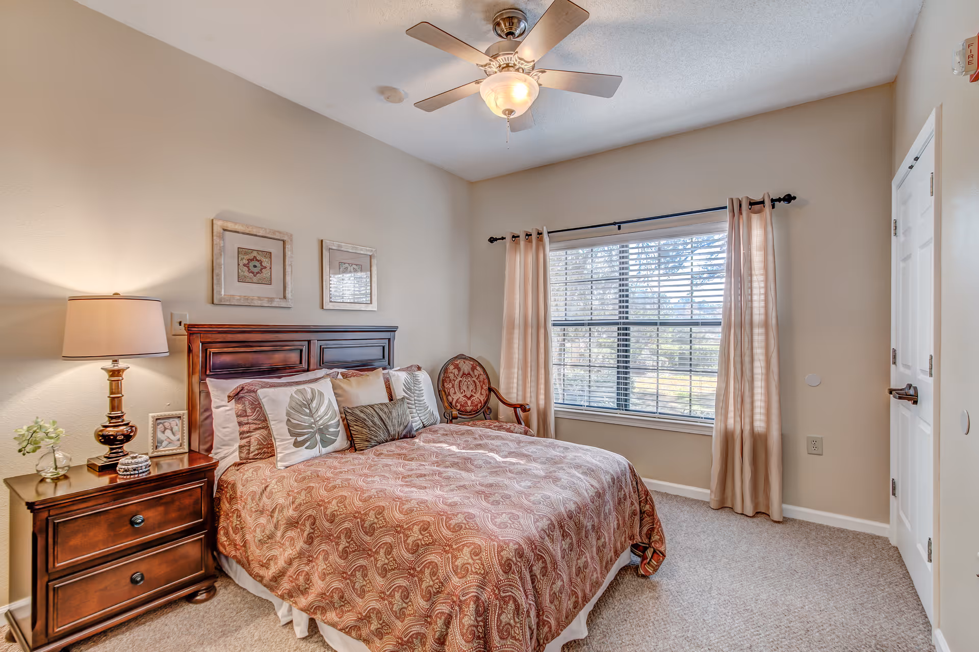 A cozy bedroom with a large window covered by beige curtains and blinds. The room features a bed with a patterned bedspread and multiple decorative pillows, a wooden headboard, a wooden nightstand with a lamp, a small plant, and a framed photo. There is also an upholstered chair with a floral pattern near the window, a ceiling fan with a light fixture, and a closed white door.