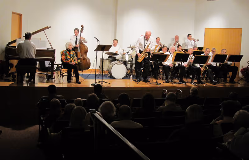 A musical band performing on a stage with various instruments including a piano, double bass, drums, saxophones, and trumpets. An elderly woman is seated on the stage near the piano. The audience is seated in front of the stage in a dimly lit room.