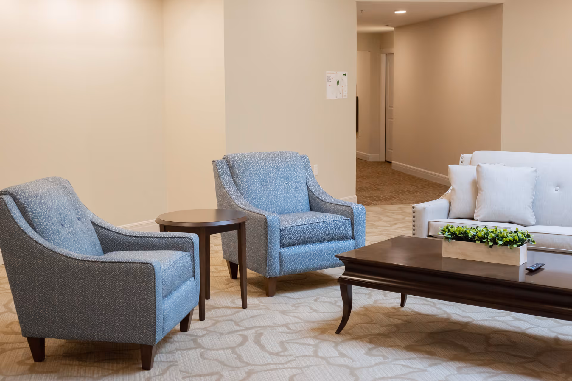 A cozy seating area in a senior living facility featuring two blue upholstered armchairs, a small round wooden side table, a white sofa with cushions, and a dark wooden coffee table with a rectangular planter of green plants on top. The room has beige walls and patterned carpet flooring, with a hallway visible in the background.
