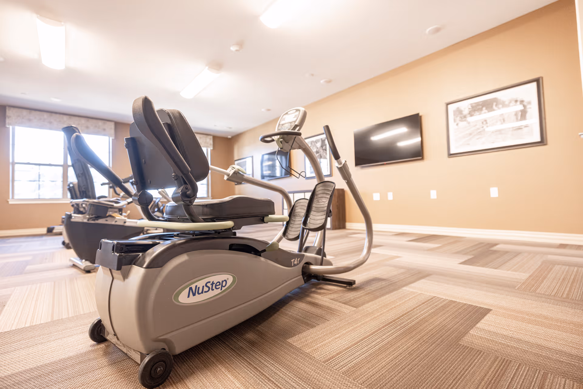 A fitness room in The Westbury Senior Living facility featuring NuStep exercise machines on a patterned carpet floor, with beige walls, framed pictures, a wall-mounted TV, and large windows letting in natural light.