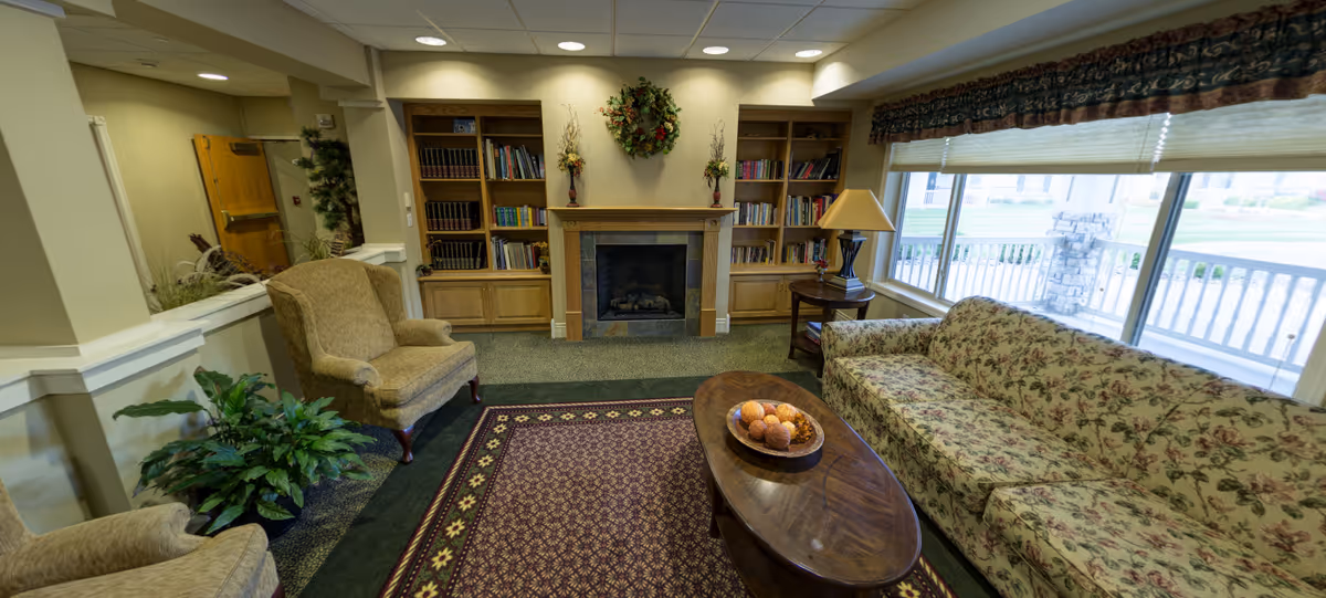 Cozy living room area with a floral patterned sofa, two beige armchairs, a wooden coffee table with decorative balls, a fireplace with a wreath above it, built-in bookshelves filled with books, a side table with a lamp, and large windows with blinds and valances letting in natural light.