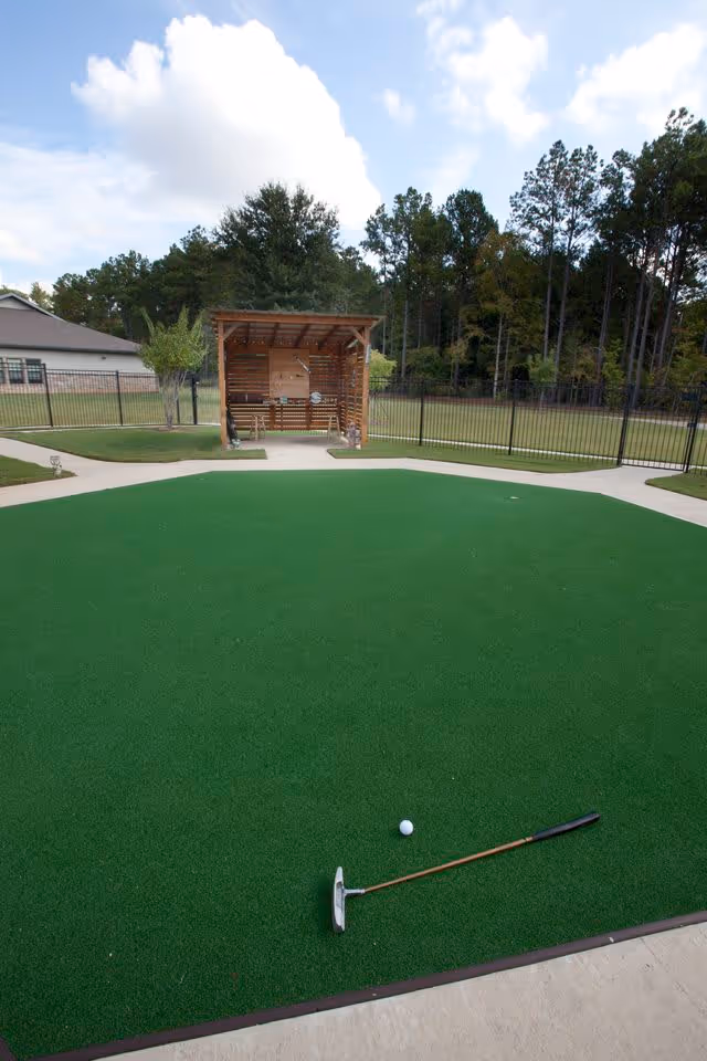 Outdoor putting green with a golf putter and golf ball on the artificial turf, surrounded by a concrete walkway. In the background, there is a wooden shelter with benches and a fenced grassy area with trees.