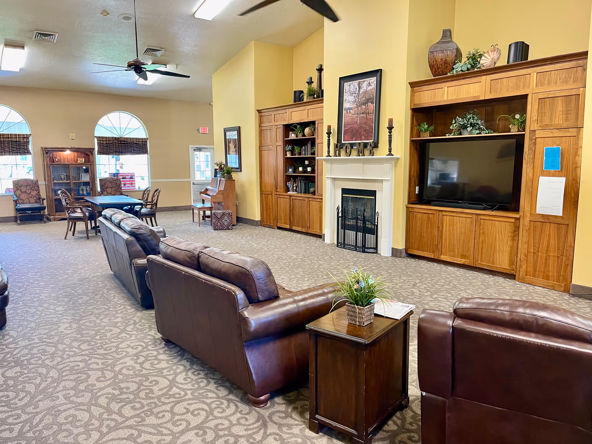 A spacious living room area in an assisted living facility featuring brown leather sofas arranged around a small wooden table with a plant. The room has a carpeted floor with a patterned design, a fireplace with decorative items on the mantel, a large flat-screen TV set within wooden cabinetry, and a piano against the far wall. There are also tables and chairs near large arched windows with blinds, ceiling fans, and warm yellow walls.