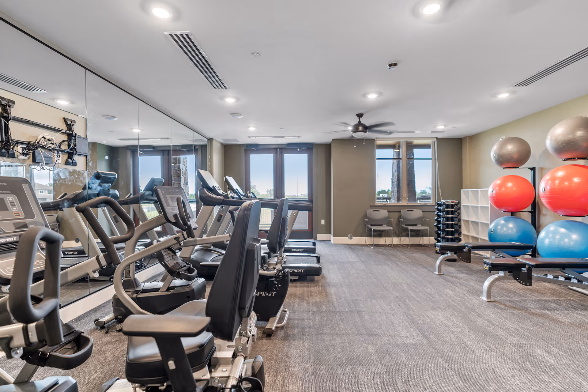 A fitness room with exercise equipment including stationary bikes and treadmills lined up facing a mirrored wall. On the right side, there are colorful exercise balls stacked on racks and a set of dumbbells. The room has large windows and a door letting in natural light, with two chairs placed near the windows.