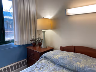 A cozy bedroom corner with a bed covered in a blue patterned quilt, a wooden nightstand with a white lamp and two small potted plants, a window with white curtains, and a wall-mounted light fixture above the bed.