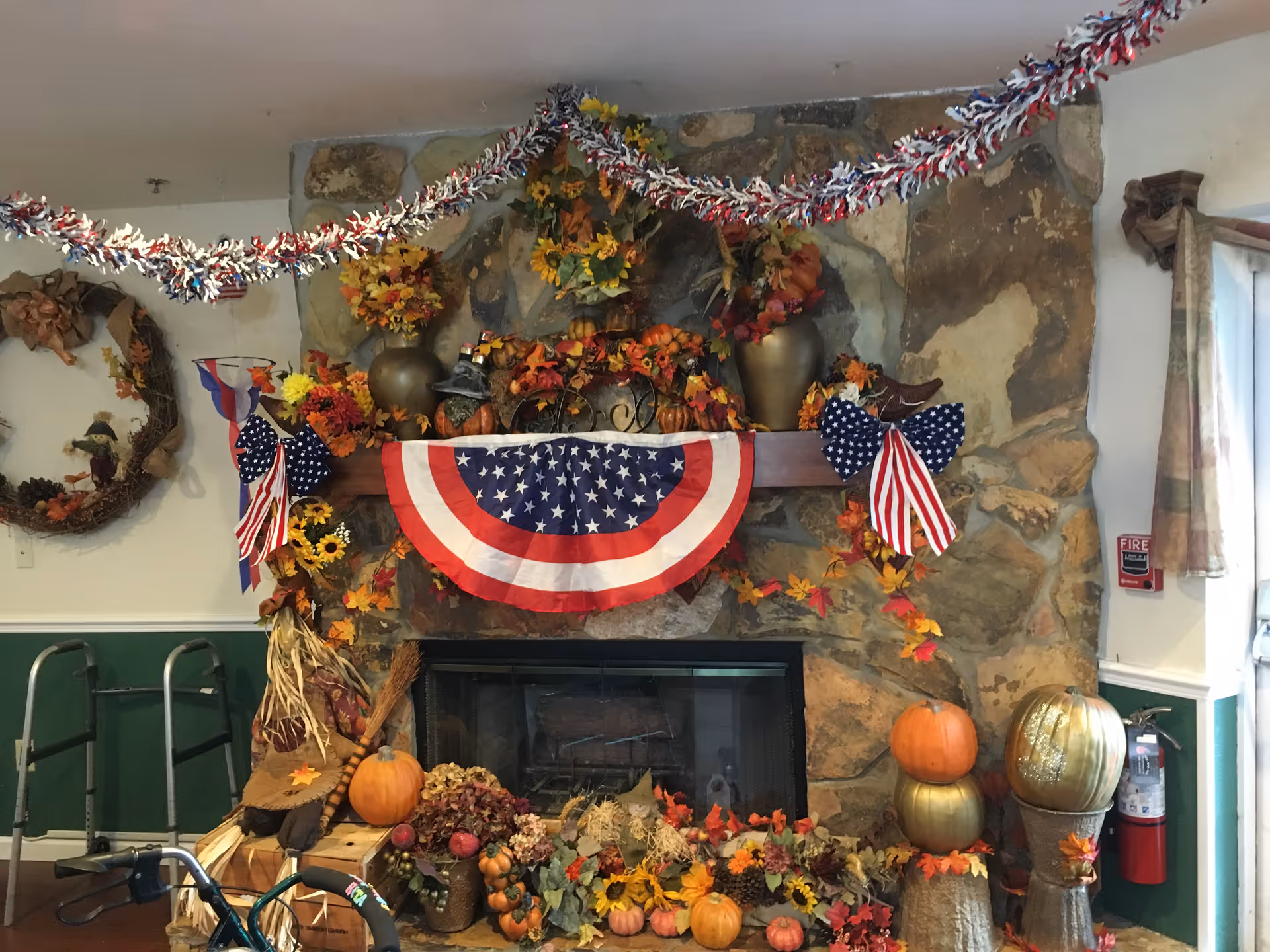 A stone fireplace decorated with autumn and patriotic decorations including pumpkins, fall leaves, flowers, and American flag-themed banners and bows. There is a scarecrow figure sitting on the left side of the hearth and a fire extinguisher mounted on the wall to the right. A walker and a wheelchair are partially visible on the left side of the image.