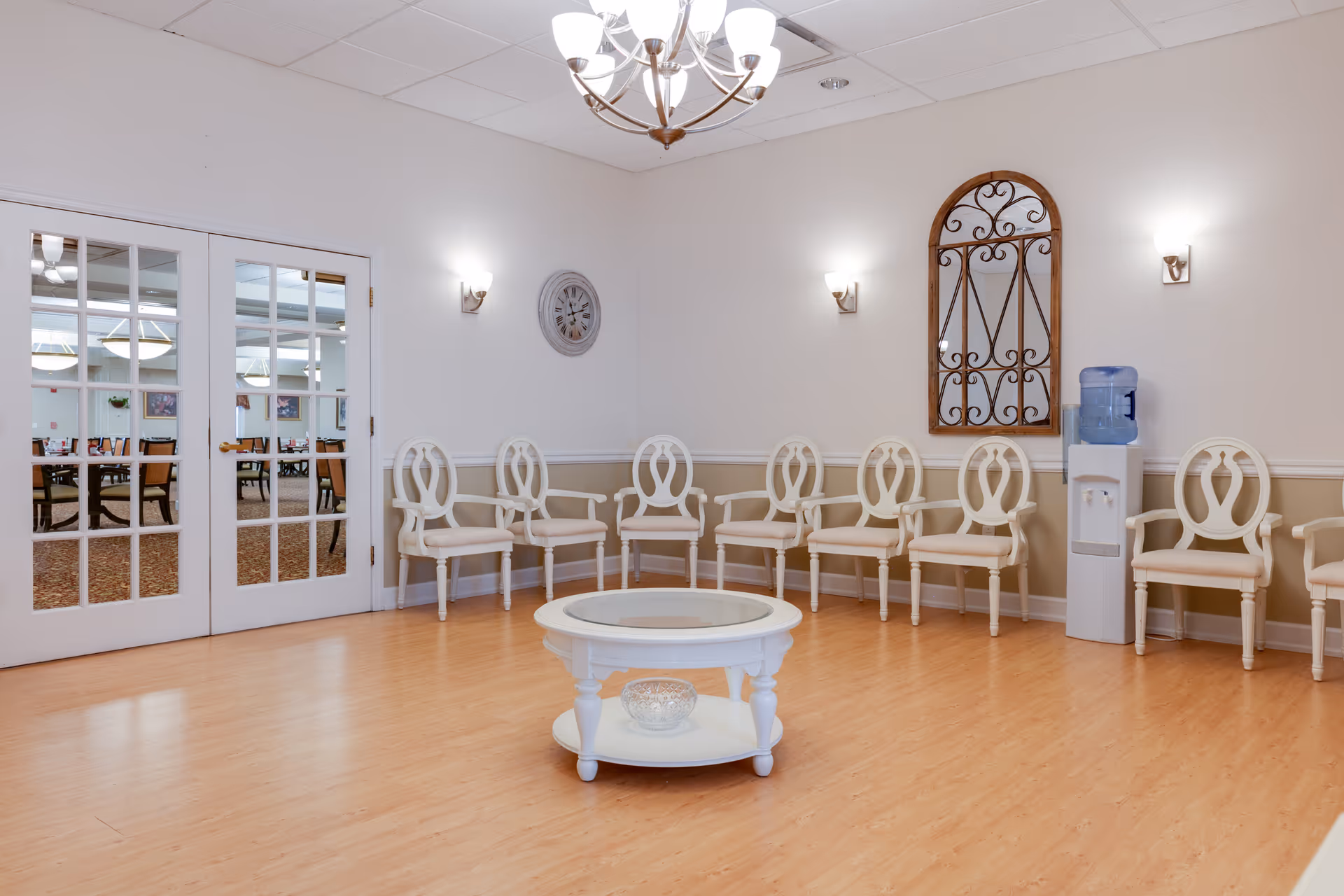 A waiting or sitting area in a senior living facility with a row of white wooden chairs with armrests arranged against the wall. The room has light-colored walls with a chair rail, a round white coffee table with a glass top in the center, a water dispenser, a decorative wall clock, and a large decorative mirror with an ornate frame. Double glass doors lead to a dining area with tables and chairs visible through the doors.