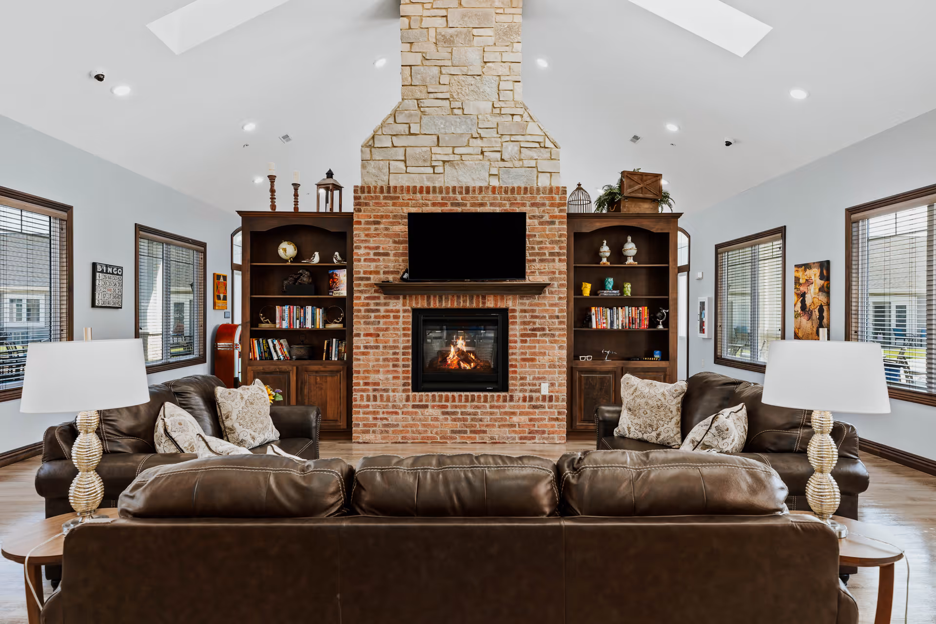 A cozy living room with a large brick and stone fireplace at the center, flanked by two wooden bookshelves filled with books and decorative items. In front of the fireplace are three brown leather sofas arranged around a coffee table, with two table lamps on side tables. The room has large windows with blinds and light-colored walls.