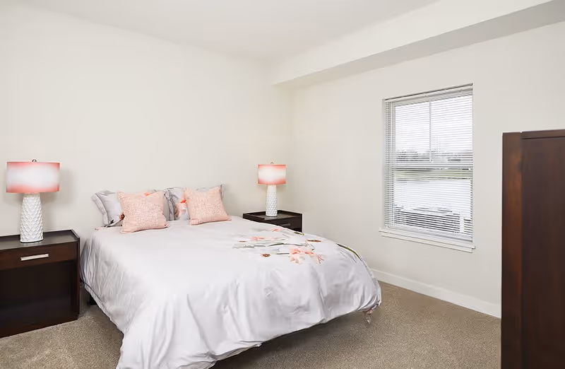 Minimal bedroom with a bed dressed in floral bedding, two bedside tables with pink-shaded lamps, and a window overlooking a body of water.