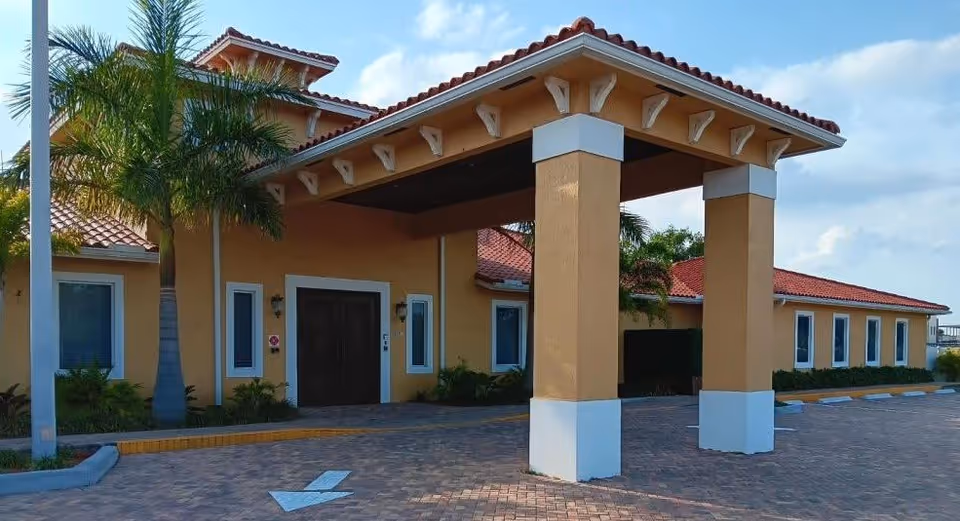 Exterior view of The Manor of Fort Pierce building with a covered entrance supported by two large columns, yellow walls, red tiled roof, palm trees, and a paved driveway with a white directional arrow.