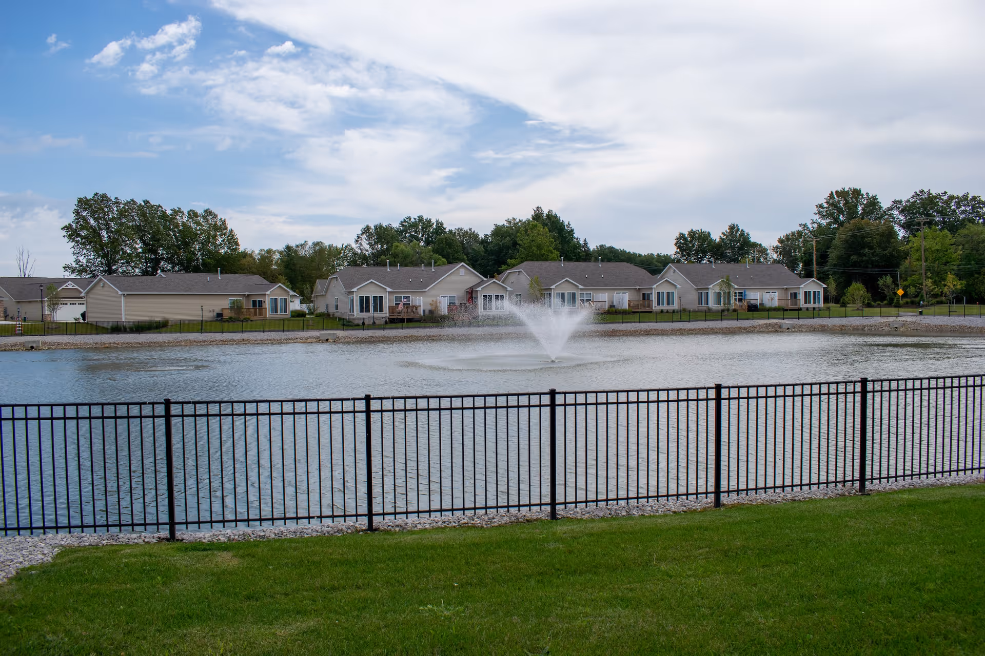 A fenced pond with a water fountain in the center, surrounded by green grass and a row of single-story residential buildings in the background under a partly cloudy sky.
