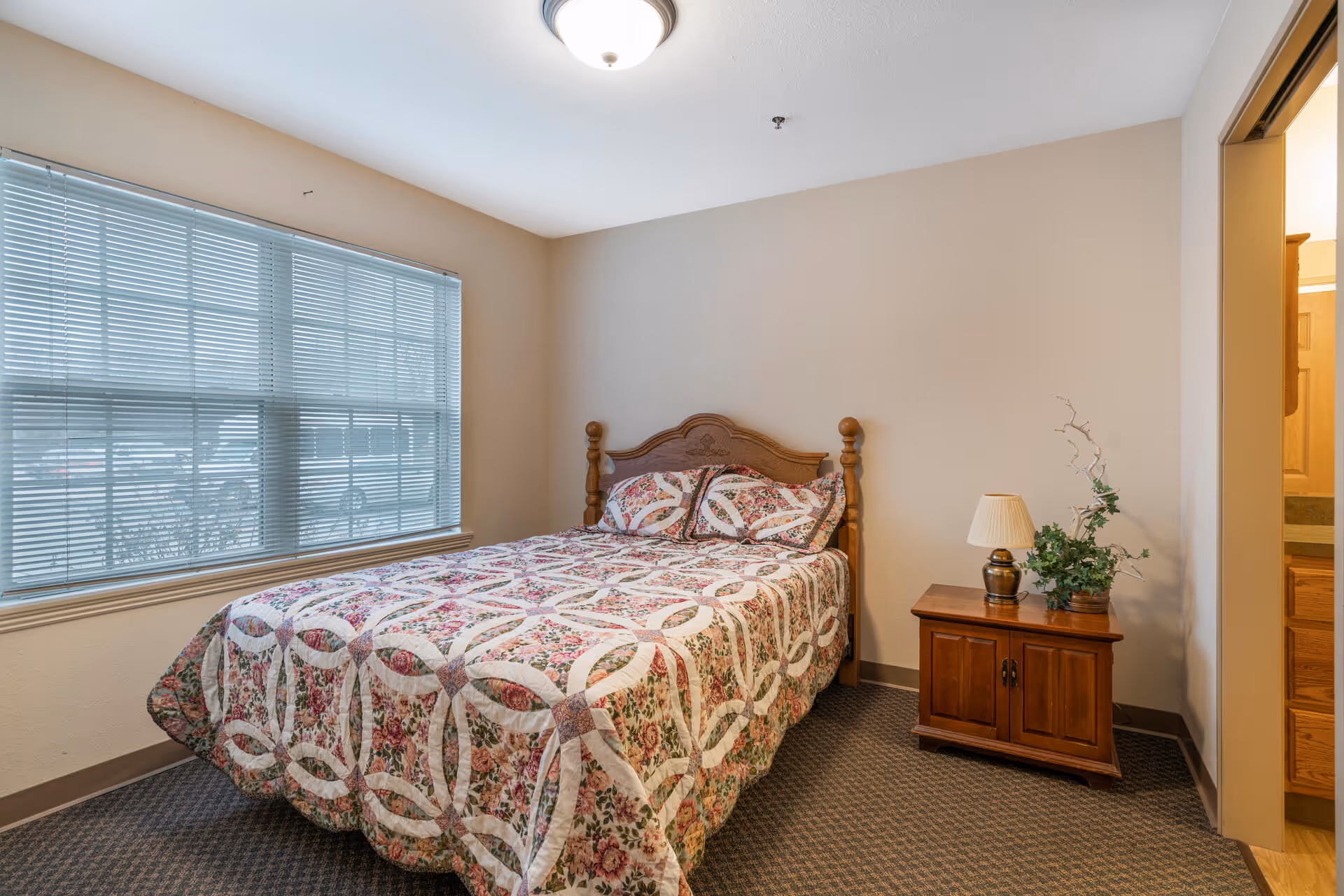 A bedroom with a wooden bed frame and a floral patterned quilt and matching pillows. There is a wooden nightstand next to the bed with a lamp and a small plant. A large window with blinds is on the left wall, and an open doorway leading to another room is on the right.