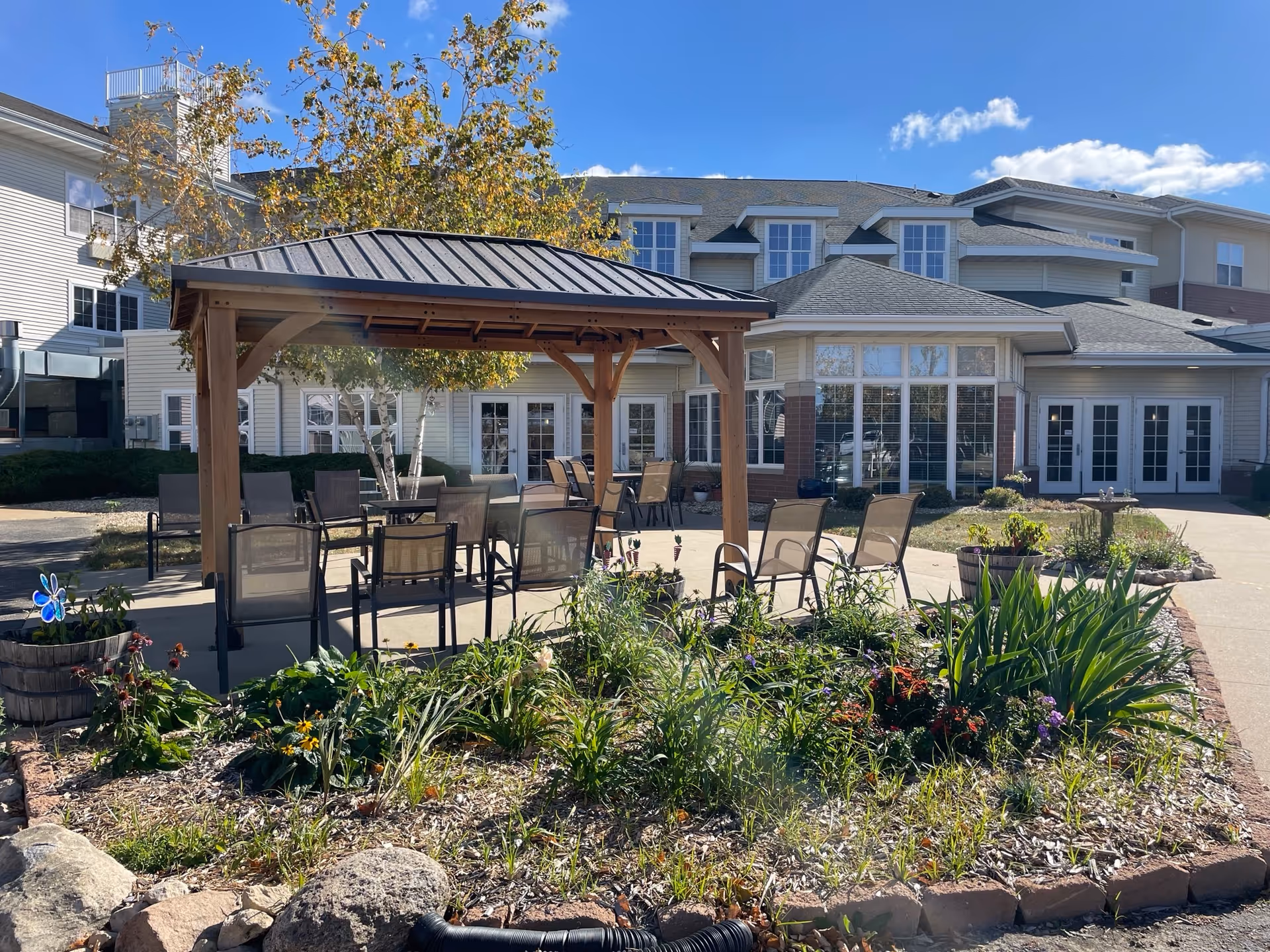 Outdoor patio area at a senior living facility with a wooden gazebo covering several chairs and tables. There is a garden bed with various plants and flowers in the foreground, and the multi-story building with large windows is visible in the background under a clear blue sky.