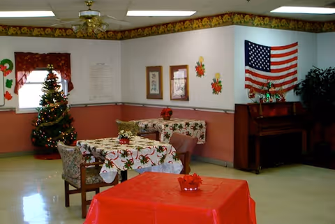Decorated common dining/activity room with tables and chairs, a piano, an American flag, and a small Christmas tree.