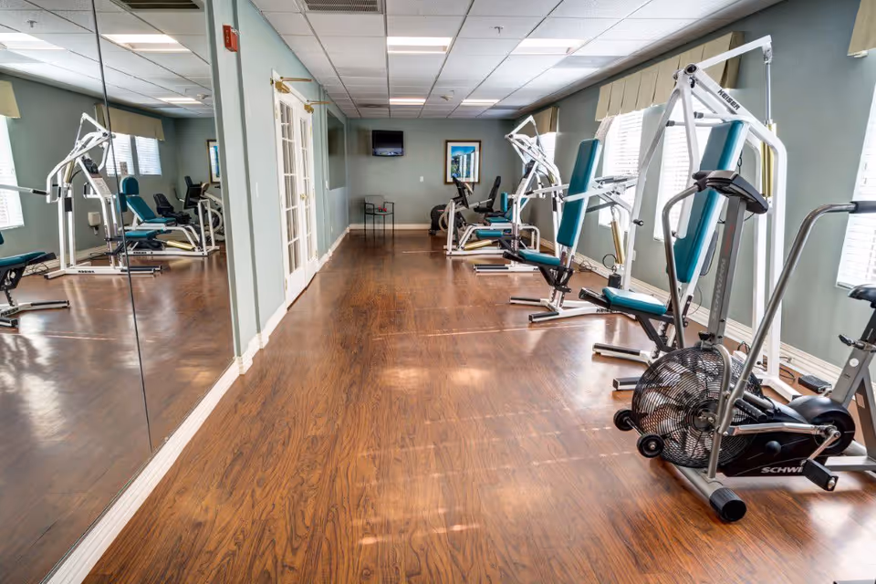 A fitness room with wooden flooring, several exercise machines including weight machines and a stationary bike, large mirrors on one wall, windows with blinds on the opposite side, and a small wall-mounted TV at the far end.