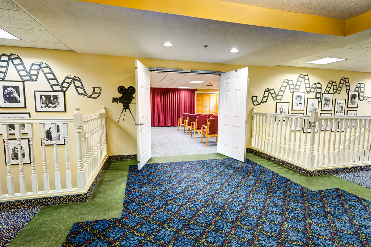 Interior hallway of a senior living facility with patterned blue and green carpet, white railings, and walls decorated with black and white movie-themed photos and film reel designs. Double white doors open to a small theater room with red chairs and red curtains.