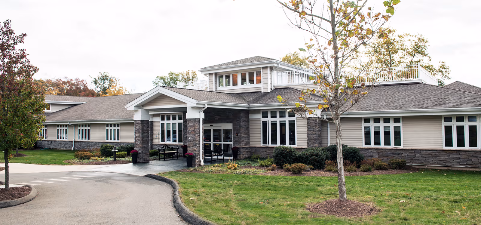 Exterior view of a single-story senior living facility building with beige siding and stone accents, large windows, a covered entrance with benches, surrounded by green grass and trees with autumn foliage.