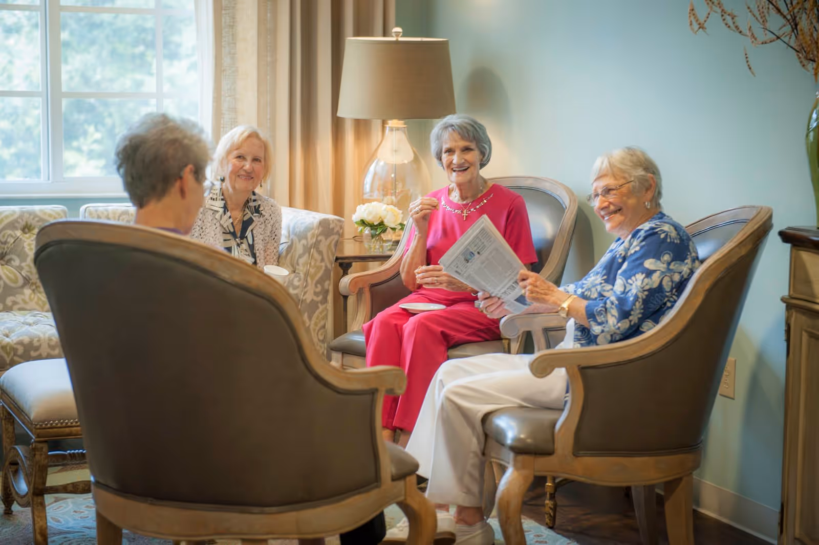 Four elderly women sitting in a cozy living room area, engaged in conversation and smiling. The room features comfortable armchairs, a sofa, a table with a lamp and flowers, and a large window with curtains letting in natural light.