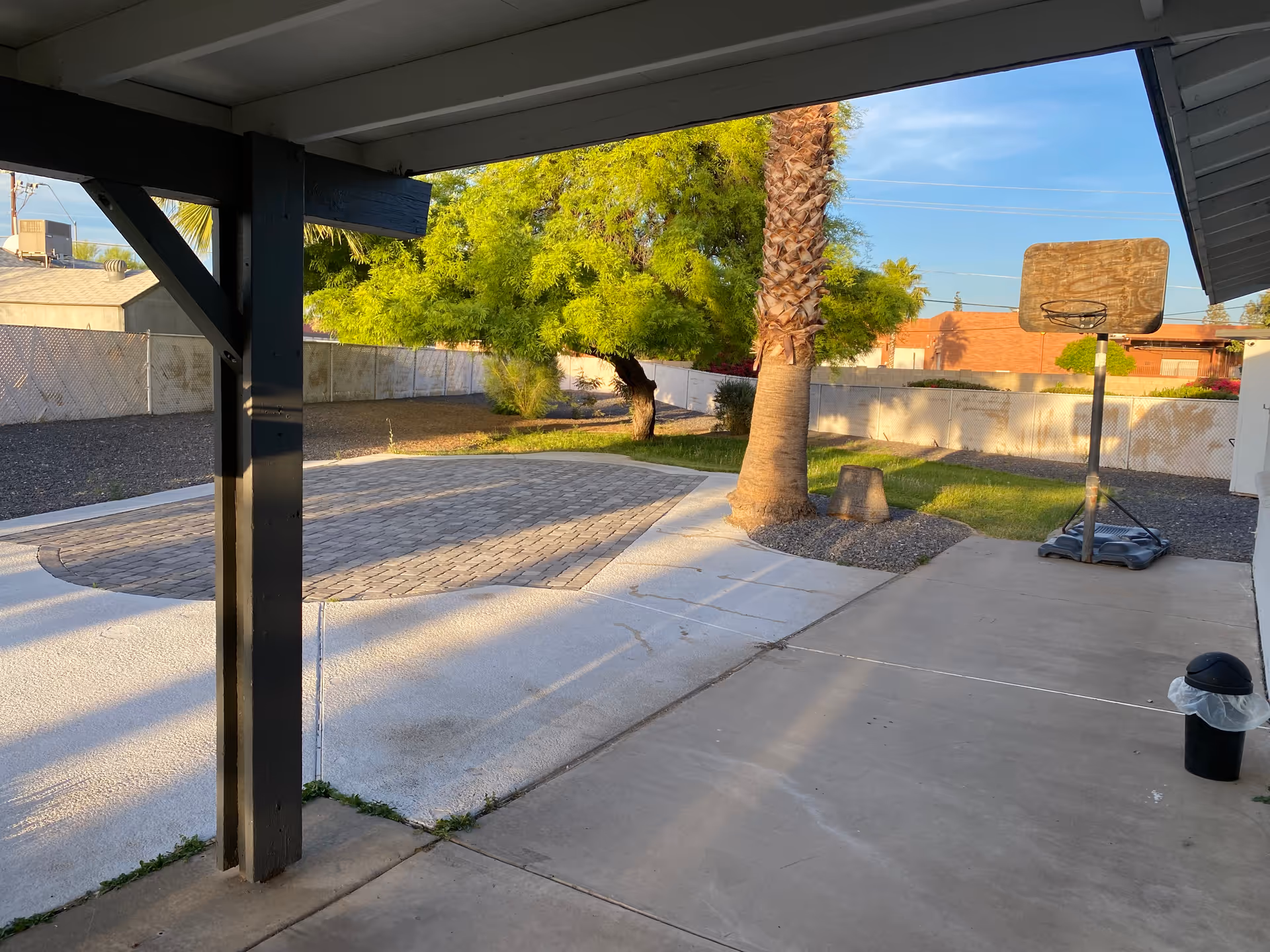 View of an outdoor patio area with a concrete floor and a small paved section. There is a palm tree and another leafy tree in the background, along with a portable basketball hoop and a trash can with a plastic liner. The area is enclosed by a chain-link fence and there are buildings visible beyond the fence under a clear blue sky.