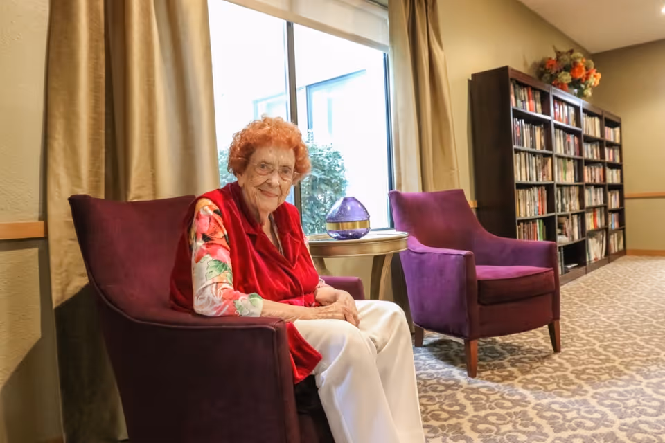 An elderly woman with curly red hair, wearing glasses, a red vest, and a floral long-sleeve shirt, sits in a purple armchair in a cozy room. Behind her is a window with beige curtains, a small round table with a decorative glass object, another purple armchair, and a tall bookshelf filled with books and topped with a flower arrangement.