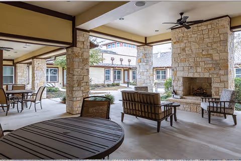 Covered outdoor patio area with stone pillars and a large stone fireplace. The space includes several seating arrangements with metal tables and chairs, and cushioned chairs around the fireplace. The patio overlooks a courtyard with greenery and buildings in the background.