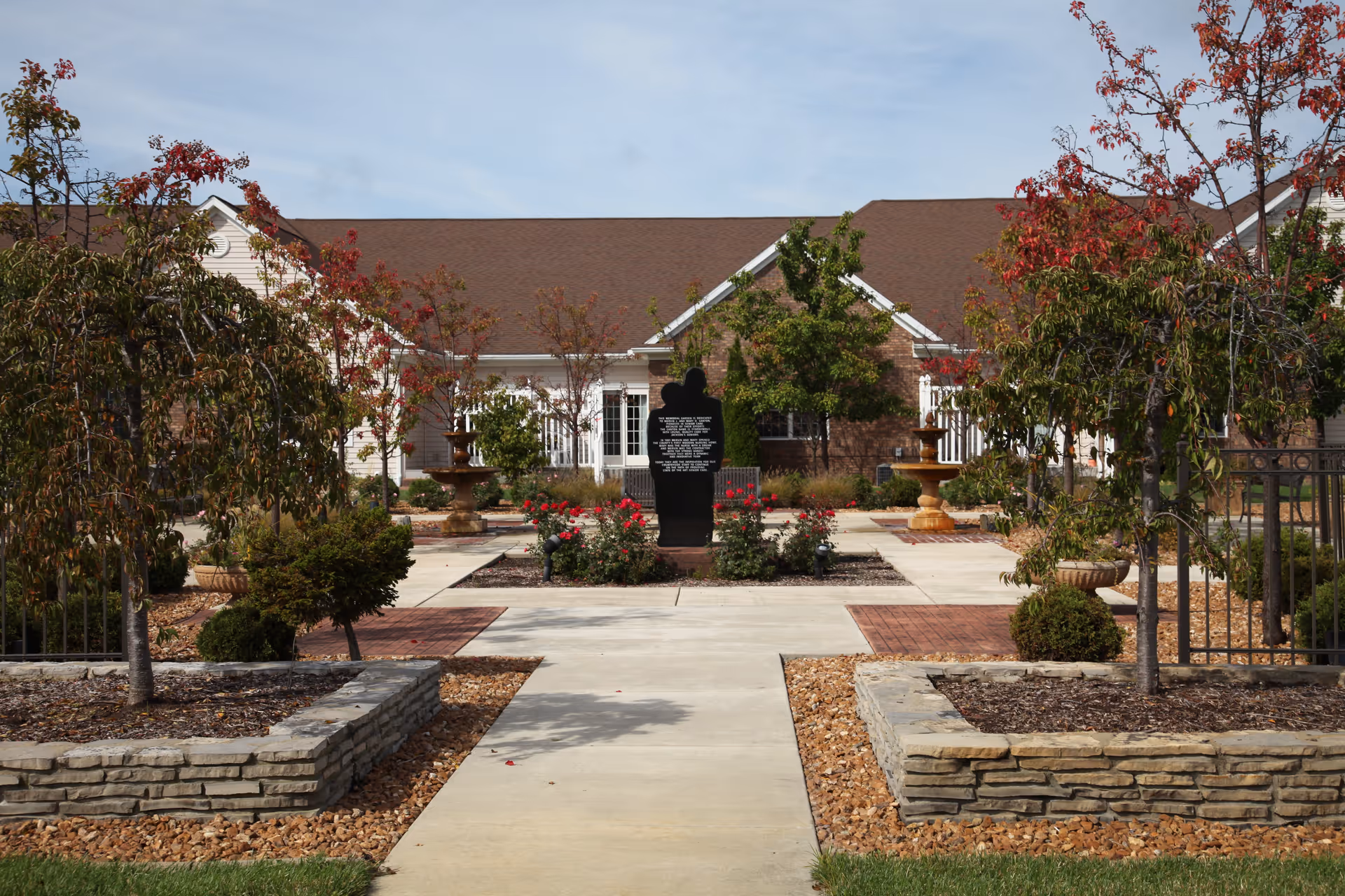 Outdoor courtyard area with a paved walkway leading to a black silhouette statue surrounded by red flowers, two fountains on either side, and trees with autumn foliage. A building with a brown roof and white siding is visible in the background under a clear blue sky.