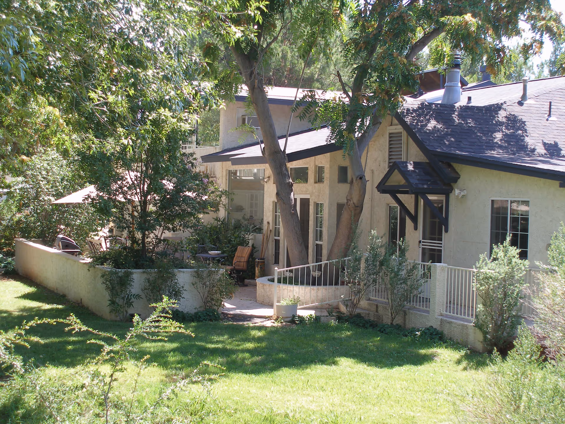 Outdoor view of a residential assisted living facility showing a patio area with chairs, surrounded by trees and greenery, and a building with beige walls and dark roof.
