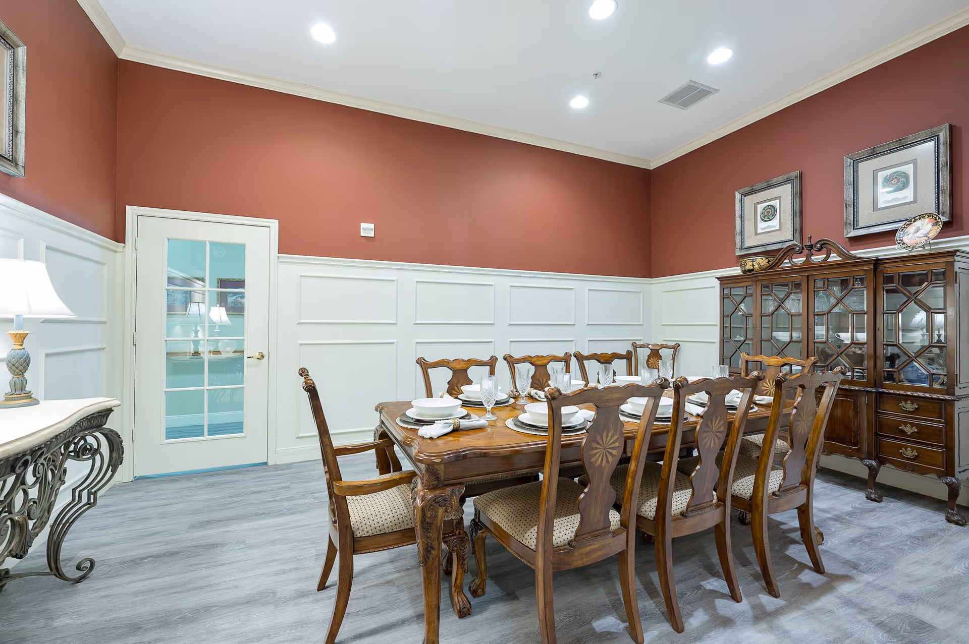 A formal dining room with a large wooden dining table set for eight people with white plates, bowls, and glasses. The room has red upper walls and white wainscoting, a glass-paneled door, a wooden china cabinet with glass doors, two framed pictures on the wall, and a decorative table with a lamp.