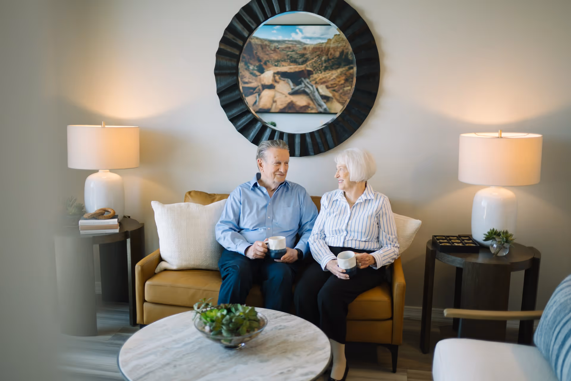 An elderly man and woman sitting on a mustard yellow couch in a cozy living room, each holding a white mug. The room features two side tables with white lamps, a round marble coffee table with a small plant centerpiece, and a large round mirror with a landscape photo above the couch.
