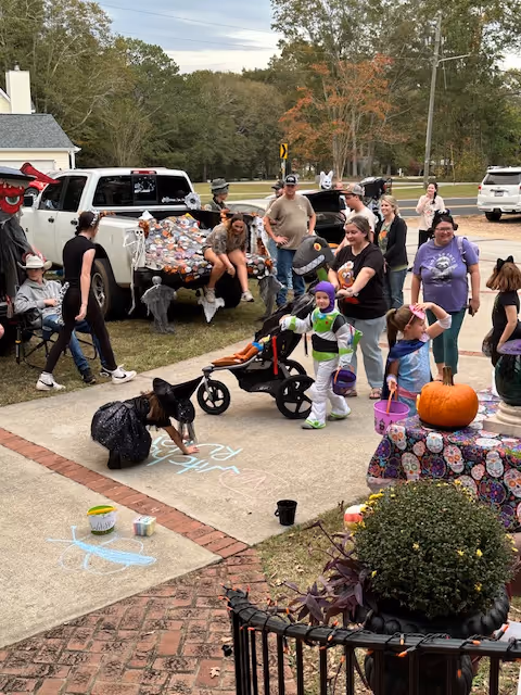 A group of children and adults dressed in Halloween costumes gathered outside near a decorated pickup truck. Children are drawing with chalk on the sidewalk, and there is a pumpkin on a table covered with a colorful cloth. Trees and parked cars are visible in the background.