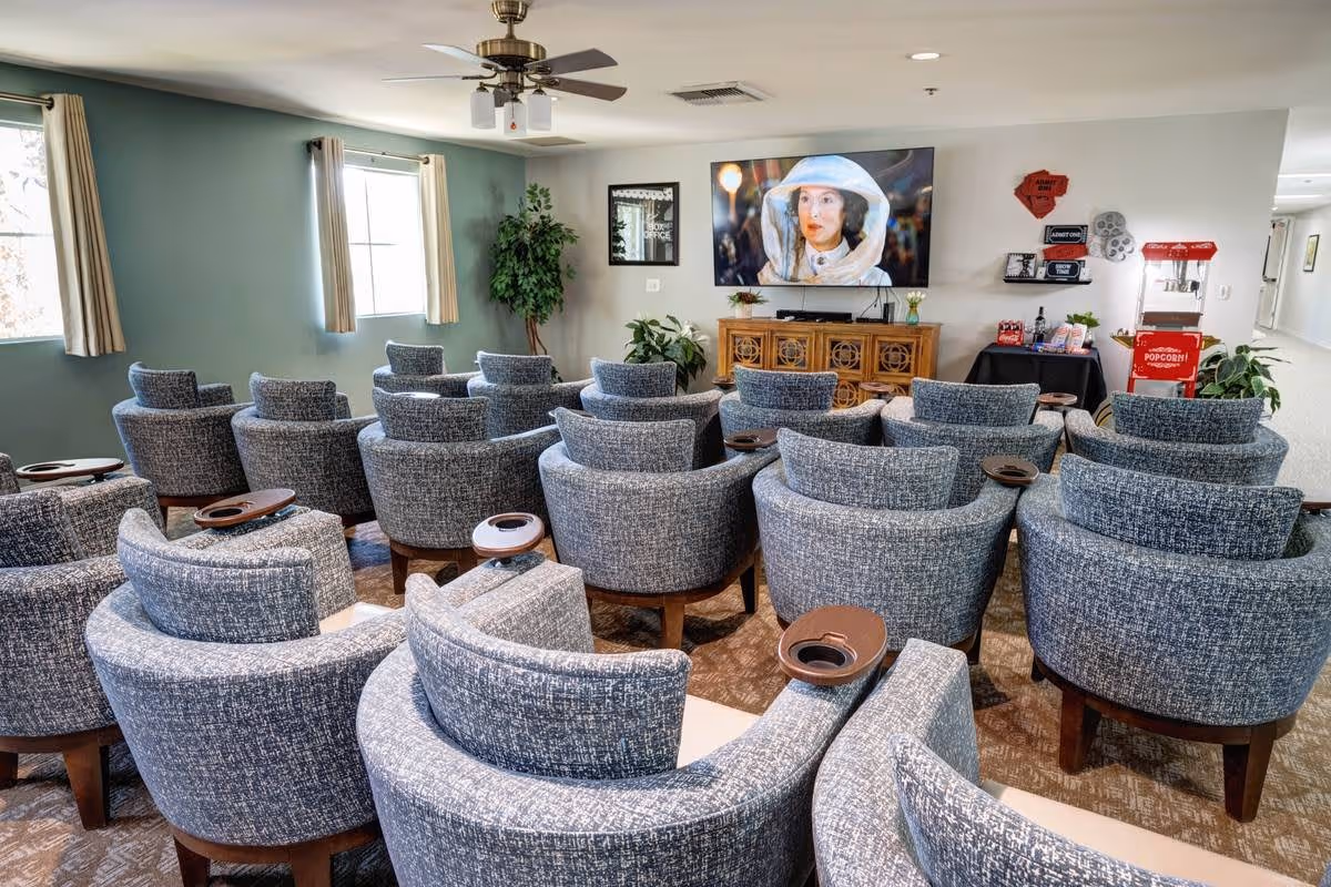 A cozy common area with multiple rows of gray upholstered armchairs facing a large flat-screen TV mounted on the wall. The TV displays a person in a white hat and outfit. The room has light green walls, two windows with beige curtains, a ceiling fan with lights, and several plants. There is a popcorn machine and a small table with snacks and drinks on the right side of the room.