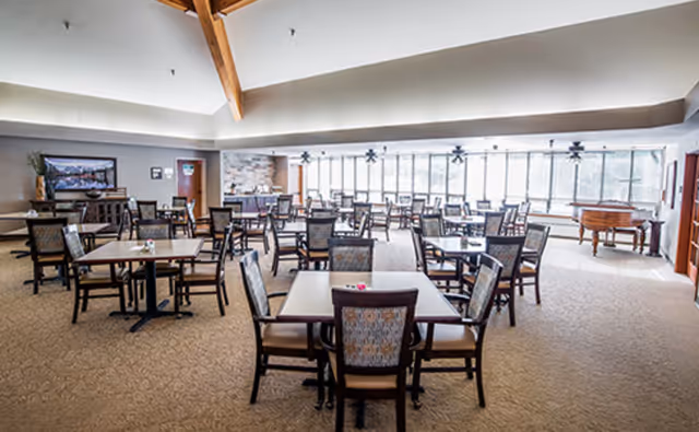 Spacious dining area with multiple square tables and upholstered chairs, large windows along the back wall and a vaulted wood-beamed ceiling.