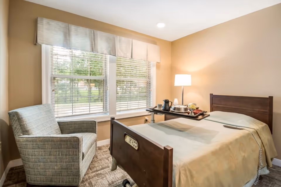 A cozy room with a single hospital-style bed covered with a beige blanket, a tray with meal items on an overbed table, a remote control on the bed, a floor lamp beside the bed, a comfortable armchair near a large window with blinds and a beige valance, and beige walls.