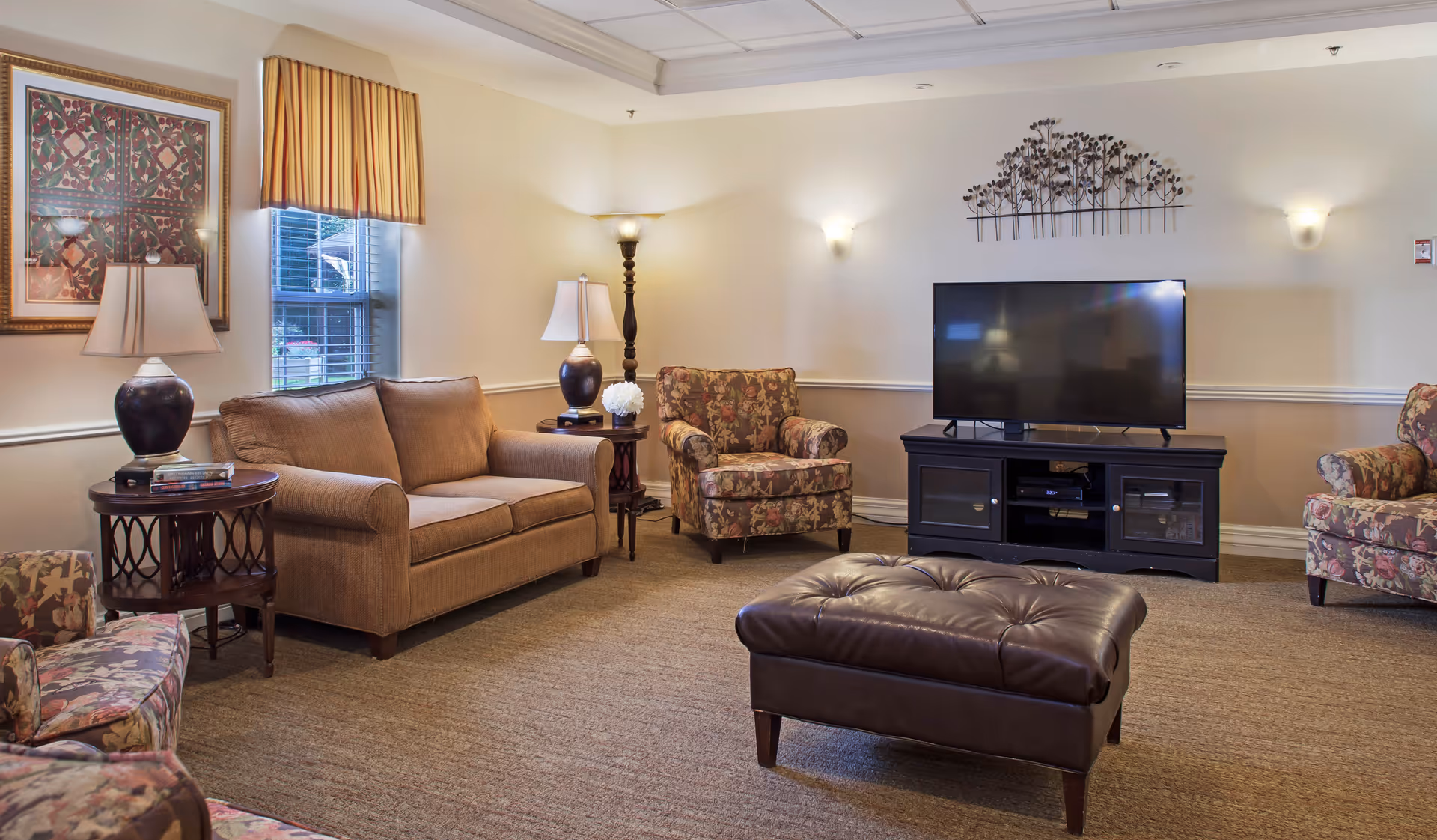 A cozy living room with a brown fabric sofa, two floral-patterned armchairs, a dark brown leather ottoman, and a black TV stand with a flat-screen television. The room has beige walls with white trim, two table lamps, a floor lamp, a window with striped valance, and decorative wall art.