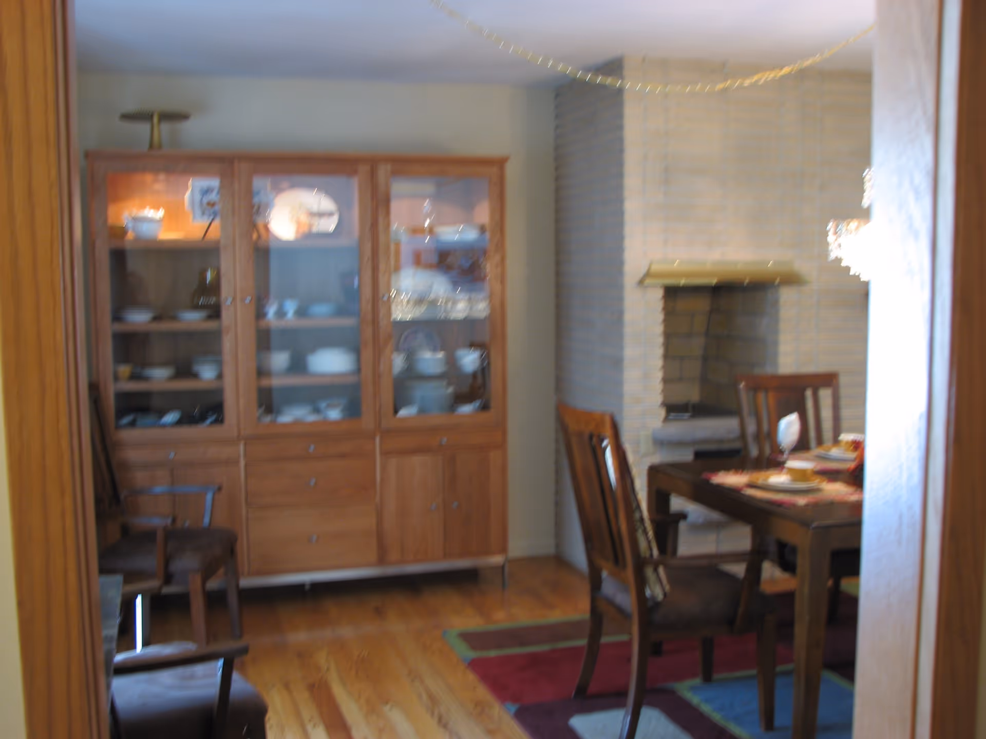 A dining room with a wooden dining table set with plates, cups, and a wine glass. There are wooden chairs around the table, a colorful rug underneath, a brick fireplace in the background, and a wooden cabinet with glass doors displaying dishes and bowls.
