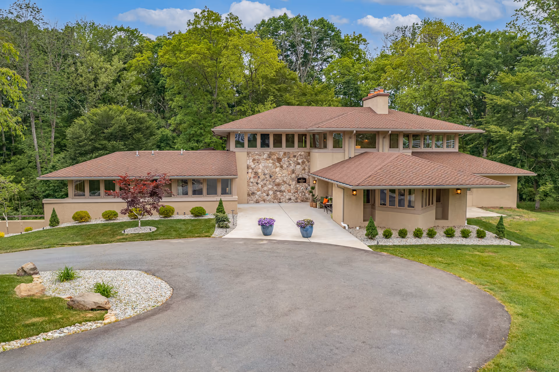 Front exterior view of a large, modern two-story building with a brown shingle roof and beige walls, surrounded by green trees and landscaping with shrubs and small trees. There is a circular driveway in front with two flower pots near the entrance.